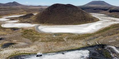 Kuppelf&ouml;rmiger dunkler H&uuml;gel in Bildmitte, umgeben von einem wei&szlig;en, ringf&ouml;rmigen Sedimentbereich auf trockenem braunem Gel&auml;nde; im Hintergrund flache Berge; im Vordergrund eine dunkle Stra&szlig;e und ein wei&szlig;es Fahrzeug; blauer Himmel mit vereinzelten Wo