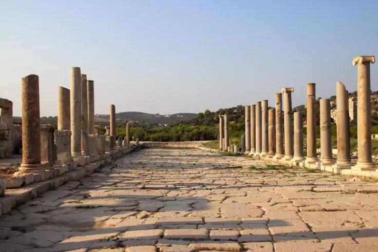 Gepflasterter Steinweg zwischen zwei Reihen steinerner S&auml;ulen, B&auml;ume und H&uuml;gel im Hintergrund, klarer Himmel