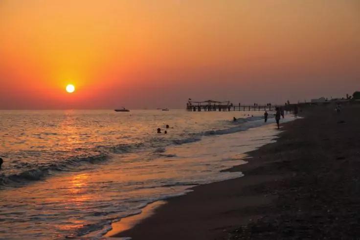 Sonnenuntergang &uuml;ber dem Meer, Sonne nahe dem Horizont, Pier rechts, Menschen am Ufer und im Wasser, Boot auf dem Meer