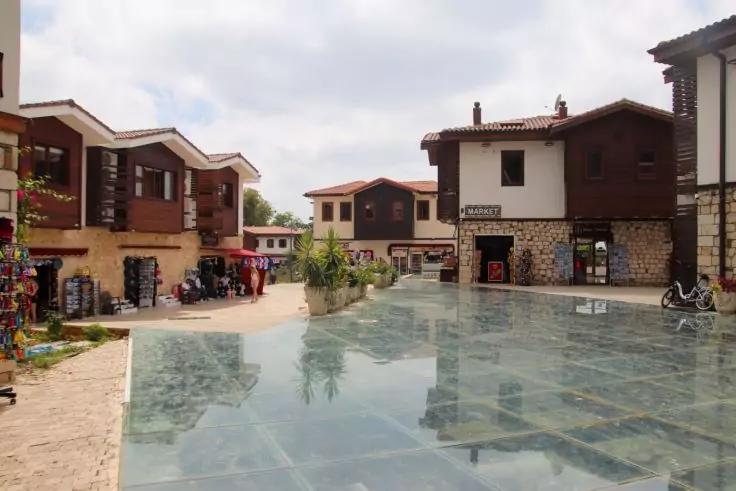 Pflasterplatz mit spiegelnder Wasserfl&auml;che, umgeben von mehrst&ouml;ckigen Geb&auml;uden mit Holz- und Steinfassaden, Schaufenstern und vereinzelten Personen unter bew&ouml;lktem Himmel