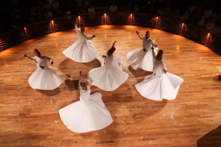 Sechs Personen in wei&szlig;en, weiten Gew&auml;ndern drehen sich mit ausgebreiteten Armen auf einer runden Holzb&uuml;hne; warme Lichter am B&uuml;hnenrand und dunkles Publikum im Hintergrund