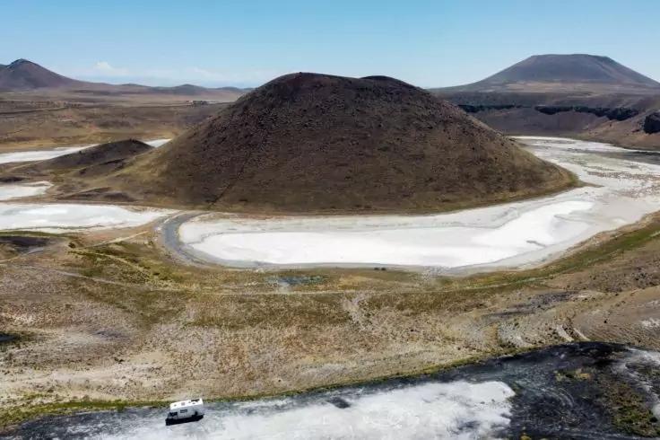 Vordergrund: unbefestigte Fl&auml;che mit einem wei&szlig;en Fahrzeug; Mitte: helle, flache Fl&auml;che um einen kuppelf&ouml;rmigen dunkelbraunen H&uuml;gel; Hintergrund: weite braune Ebenen und niedrige Berge unter blauem Himmel