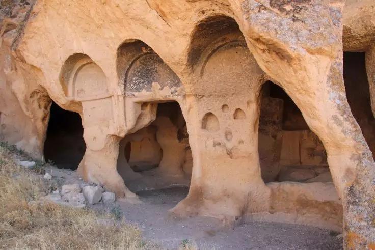 Felswand mit mehreren bogenf&ouml;rmigen &Ouml;ffnungen und Fels&shy;s&auml;ulen, sandiger Boden und trockenes Gras im Vordergrund