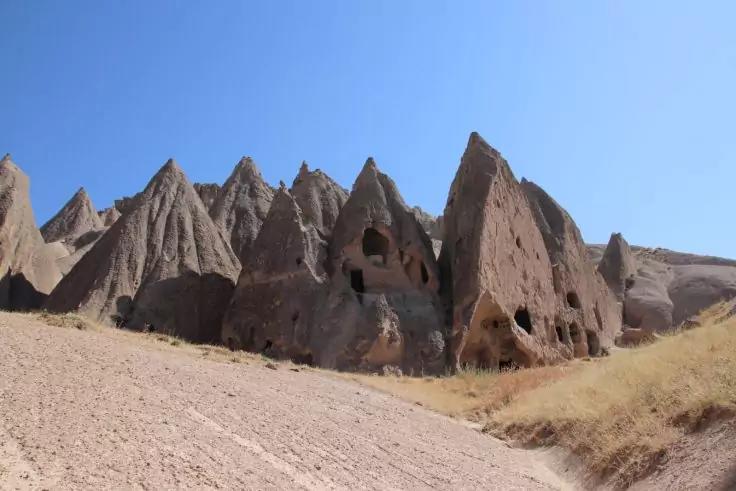Felsformationen mit mehreren &Ouml;ffnungen in den Felsw&auml;nden, sandiger Boden mit trockenem Gras und klarer blauer Himmel