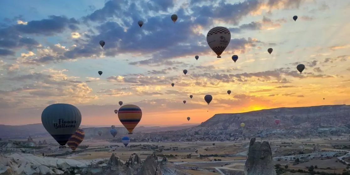 Mehrere Hei&szlig;luftballons am Himmel &uuml;ber einer felsigen Landschaft mit Wolken und orangefarbenem Horizont