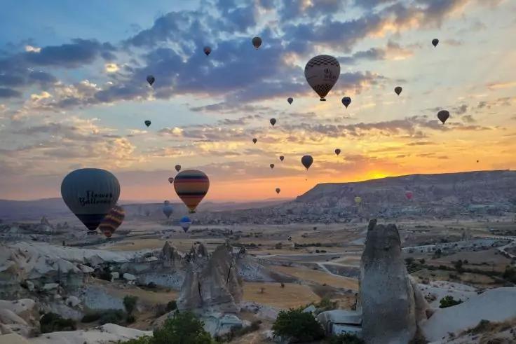 Mehrere Hei&szlig;luftballons &uuml;ber einem felsigen Tal, bunter Himmel mit Sonne am Horizont