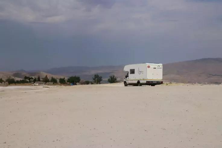 Wei&szlig;es Wohnmobil auf weiter, heller sandiger Fl&auml;che; vereinzelte B&auml;ume und niedrige Berge im Hintergrund unter bew&ouml;lktem Himmel