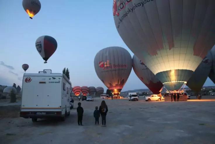 Mehrere aufgeblasene Hei&szlig;luftballons auf einem offenen Platz, einige mit brennenden Brennern, ein wei&szlig;es Wohnmobil und drei Personen im Vordergrund