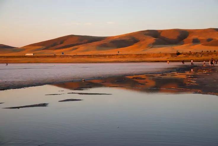 Flache Wasserfl&auml;che mit Spiegelung, sandige D&uuml;nen im Hintergrund, wenige Menschen am rechten Ufer, klarer Himmel