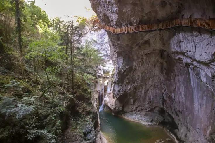 Schmale Felsenschlucht mit h&ouml;lzernem Steg an der rechten Felswand, gr&uuml;nem Wasserbecken und kleinem Wasserfall, links B&auml;ume und Vegetation