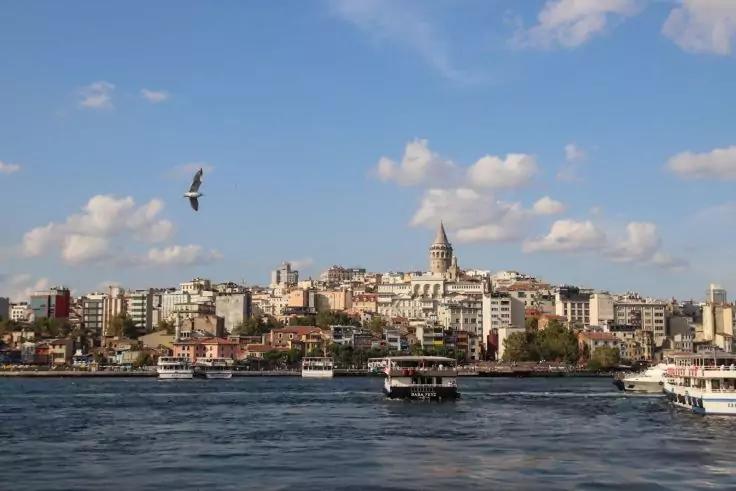 Stadtansicht am Wasser mit dicht stehenden Geb&auml;uden, einem hohen Turm in der Mitte, mehreren F&auml;hren und Booten auf dem Wasser und blauem Himmel mit vereinzelten Wolken