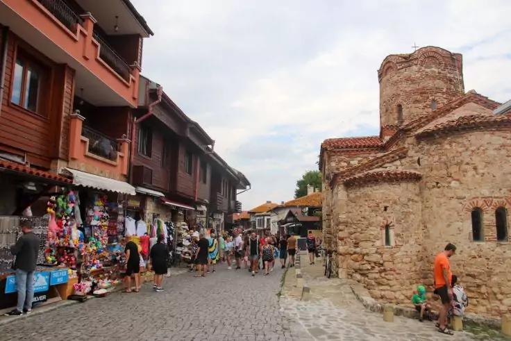 Gepflasterte Stra&szlig;e mit Marktst&auml;nden und Menschen, links h&ouml;lzerne zweist&ouml;ckige Geb&auml;ude, rechts steinernes Geb&auml;ude mit rundem Turm, bew&ouml;lkter Himmel