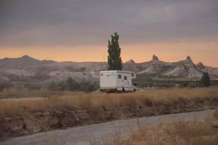 Wohnmobil auf einem Feldweg vor felsiger Landschaft, vereinzelte B&auml;ume und orange-grauer Himmel