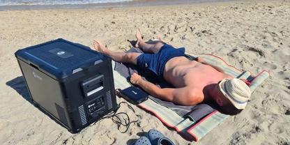 Person liegt auf dem Bauch auf gestreifter Strandmatte, tr&auml;gt Strohhut und blaue Badehose; links ein gro&szlig;es dunkles rechteckiges Ger&auml;t mit Kabel; graue Sandalen daneben; Sand und Meer im Hintergrund