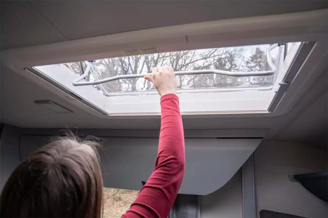 Person von hinten, die mit ausgestrecktem Arm ein Dachfenster öffnet; Bäume durch das Fenster sichtbar