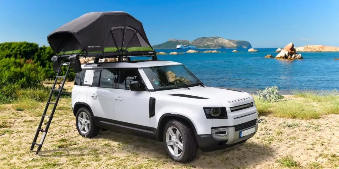 Wei&szlig;es SUV mit aufgestelltem Dachzelt und Leiter, geparkt an grasigem Strand; Meer und Felsen im Hintergrund