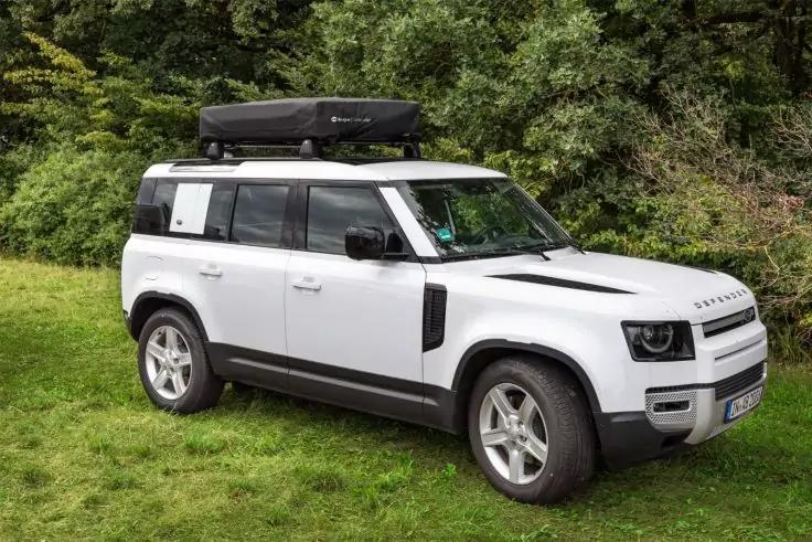 Weißer Geländewagen mit schwarzer Dachbox, drei-viertel-Ansicht von vorn, auf Gras vor Bäumen