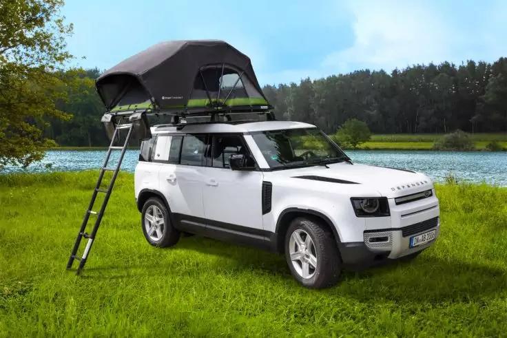 Weißer Geländewagen mit ausgeklapptem Dachzelt und Leiter, geparkt auf einer Wiese am See, Bäume im Hintergrund