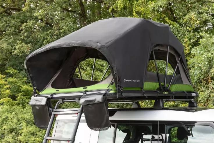 Aufgeklapptes schwarzes Dachzelt mit grünem Boden auf dem weißen Fahrzeugdach, Metall-Gepäckträger, Leiter und zwei Seitentaschen, Bäume im Hintergrund