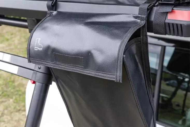 Schwarze glänzende Tasche mit Klappe, an einem schwarzen Metallträger mit Gurten befestigt; kleines weißes Symbol auf der Klappe; im Hintergrund Autofenster und Gras