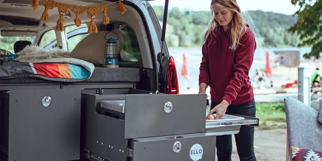 Person steht am offenen Heck eines Campingbusses und zieht eine ausziehbare Schublade heraus; im Bus liegen Bettzeug und Gep&auml;ck, im Hintergrund ein See mit Sonnenschirmen