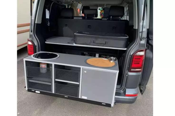 Offener Kofferraum eines dunklen Vans mit herausgezogenem grauem M&ouml;belmodul: links rundes Ausschnitt, mittig zwei offene F&auml;cher, rechts graue T&uuml;r, oben Ablage mit schwarzer Kunststoffschale, hinten zwei Kopfst&uuml;tzen