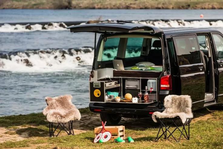 Schwarzer Van mit ge&ouml;ffneter Heckklappe und sichtbarer Heckk&uuml;che, zwei klappbare St&uuml;hle mit Fell&uuml;berz&uuml;gen, Holzkiste und Picknickgeschirr auf Gras am Fluss mit kleinen Wasserf&auml;llen im Hintergrund