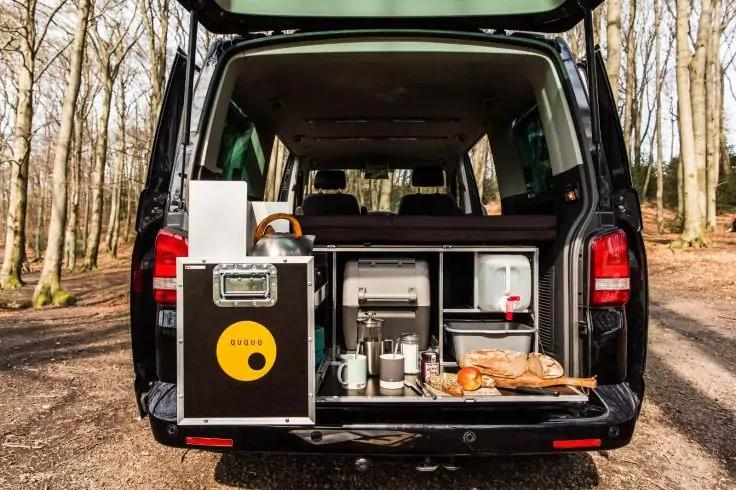 Offenes Heck eines schwarzen Vans mit Aufbewahrungskisten und Regalf&auml;chern, sichtbare Kaffeemaschine, Tassen und Brot auf einem Holzbrett; im Hintergrund Wald mit d&uuml;nnen B&auml;umen