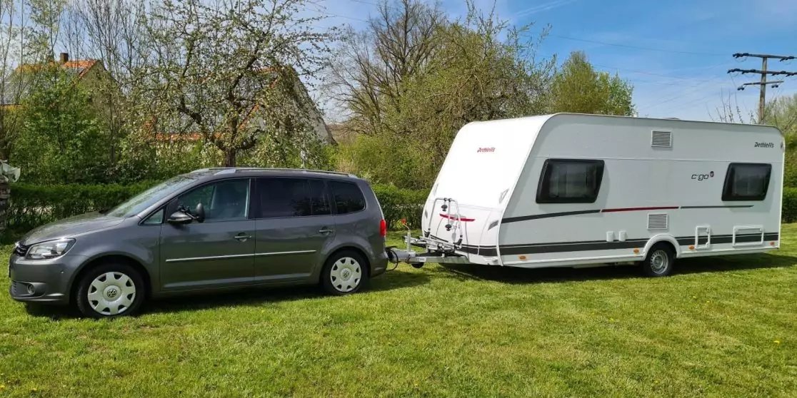 Graues Auto mit angeh&auml;ngtem wei&szlig;em Wohnwagen auf gr&uuml;ner Wiese, B&auml;ume und blauer Himmel im Hintergrund