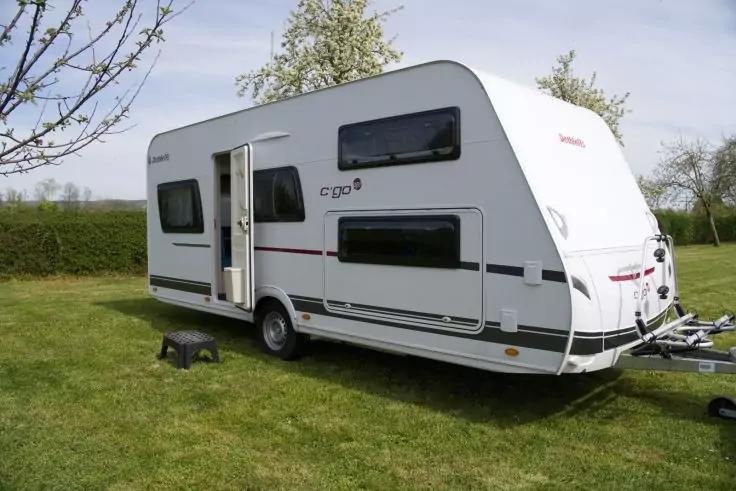 Wei&szlig;er Wohnwagen auf einer Grasfl&auml;che neben B&auml;umen, offene T&uuml;r, kleiner schwarzer Tritt neben der T&uuml;r, sichtbare Deichsel und Fenster