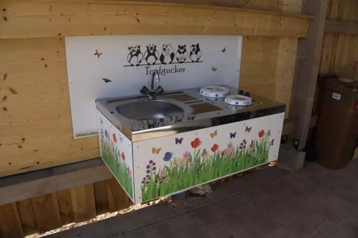 Wandmontiertes Metallwaschbecken mit Wasserhahn und zwei wei&szlig;en Schalen rechts; Vorderseite bemalt mit Gras, Blumen und Schmetterlingen; wei&szlig;es R&uuml;ckbrett mit vier Eulenzeichnungen und dem Schriftzug "Trogucker"; Holzwand und braune M&uuml;lltonne rechts