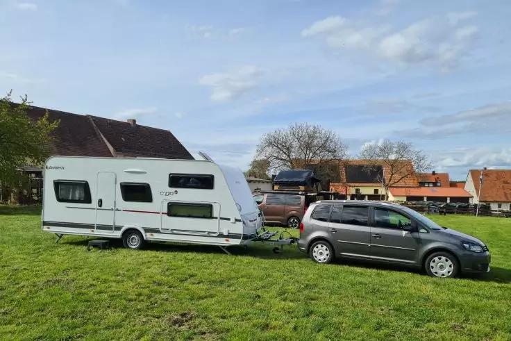 Graues Auto mit angeh&auml;ngtem wei&szlig;em Wohnwagen auf gr&uuml;ner Wiese, im Hintergrund H&auml;user, B&auml;ume und teilweise bew&ouml;lkter Himmel