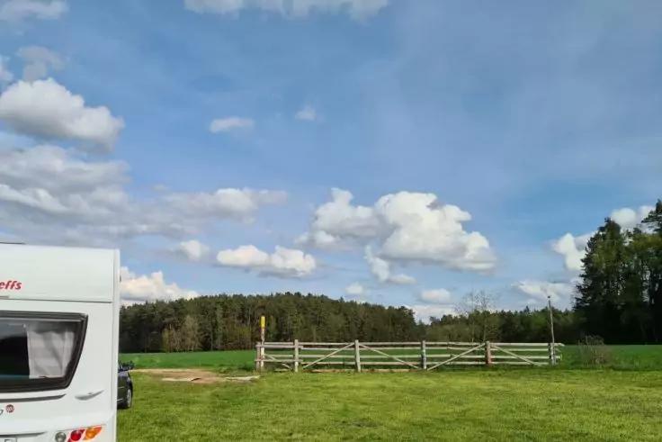 Teilansicht eines wei&szlig;en Wohnmobils links, gr&uuml;ner Rasen, h&ouml;lzerner Zaun, Wald im Hintergrund, blauer Himmel mit Wolken