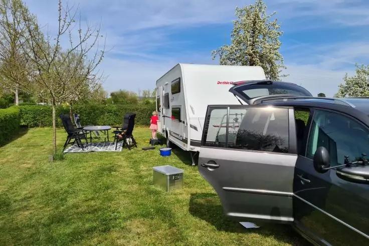 Wohnwagen auf einer Wiese neben einem Auto mit ge&ouml;ffneter T&uuml;r; davor Tisch und St&uuml;hle auf einer Decke, eine K&uuml;hlbox und ein kleiner Hocker; zwei Personen neben dem Wohnwagen; bl&uuml;hende B&auml;ume und blauer Himmel mit Wolken