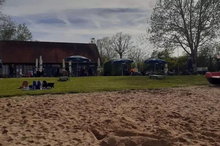 Sandfl&auml;che im Vordergrund, Rasen mit sitzenden Personen, Geb&auml;ude mit rotem Satteldach, Sonnenschirme, B&auml;ume, bew&ouml;lkter Himmel