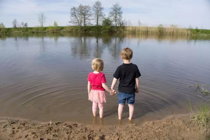 Zwei Kinder stehen im flachen Wasser, R&uuml;cken zur Kamera, halten H&auml;ndchen; sandiger Uferbereich im Vordergrund, Schilf und B&auml;ume am gegen&uuml;berliegenden Ufer, bew&ouml;lkter Himmel