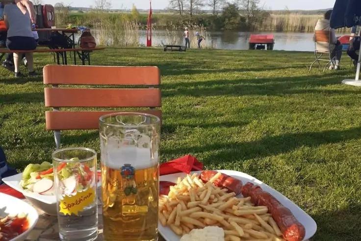 Bierglas, Wasserglas mit Zitronenscheibe, Teller mit Pommes, Wurst und Mayonnaise, kleine Salatportion, rotes Servier- oder Taschentuch auf dem Tisch, Tische und St&uuml;hle auf Rasen, sitzende Personen, See mit Schilf im Hintergrund