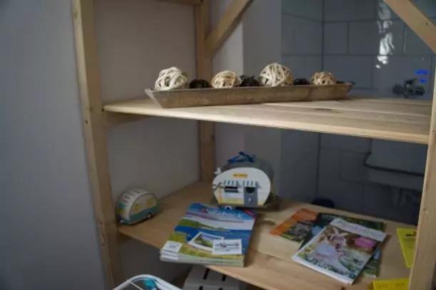 Helles Holzregal mit drei offenen B&ouml;den. Auf dem oberen Brett eine l&auml;ngliche Schale mit dekorativen, korbartigen Kugeln. Auf den unteren B&ouml;den verstreute Brosch&uuml;ren, Prospekte und zwei kleine gestreifte Dekoobjekte