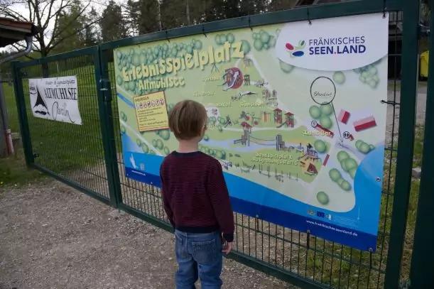 Person von hinten in dunkelrotem Pullover und blauer Jeans steht vor einer gro&szlig;en, farbigen Karte auf einem gr&uuml;nen Metallzaun; Wiese und B&auml;ume im Hintergrund