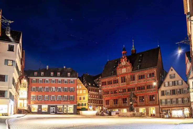 Gepflasterter Marktplatz bei Nacht, umgeben von mehreren mehrst&ouml;ckigen Giebelgeb&auml;uden mit verzierten Fassaden und beleuchteten Schaufenstern, dunkler blauer Himmel