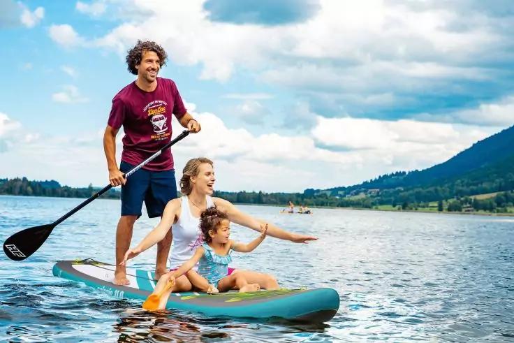 Drei Personen auf einem Stand-up-Paddleboard auf einem See. Ein Mann steht mit Paddel, eine Frau sitzt, ein Kind sitzt vorn. Berge und Wolken im Hintergrund