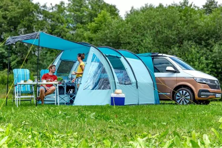 Auto mit angebautem Vorzelt; zwei Personen sitzen an einem Campingtisch mit St&uuml;hlen, K&uuml;hlbox und Ausr&uuml;stung auf einer Wiese vor B&auml;umen