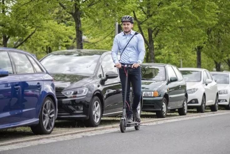 Person mit Helm f&auml;hrt auf einem E‑Scooter auf einer Stra&szlig;e neben geparkten Autos, B&auml;ume im Hintergrund