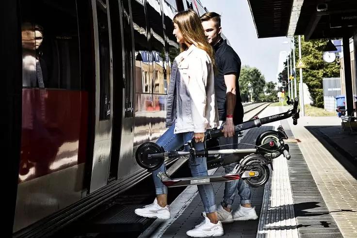 Zwei Personen (Frau vorn, Mann dahinter) mit zusammengeklappten E‑Scootern stehen an ge&ouml;ffneten Zugt&uuml;ren auf einem Bahnsteig