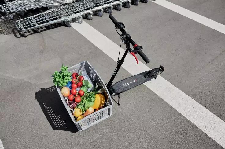 Schwarzer E-Scooter neben einem grauen Einkaufskorb mit Tomaten, Paprika, Bananen, Salat und Kr&auml;utern auf Asphalt mit wei&szlig;en Markierungen; Einkaufswagen im Hintergrund