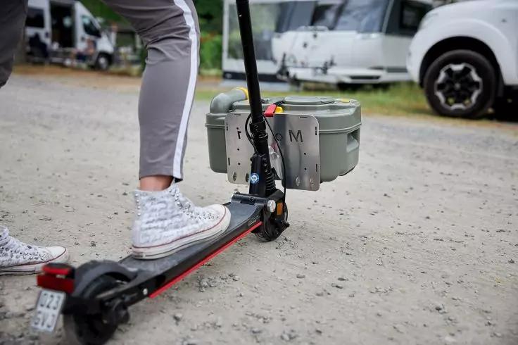 Unterbeine mit wei&szlig;en gemusterten Sneakern auf einem Elektroroller mit befestigter grauer Kunststoffbox, Kiesweg, Wohnmobile im Hintergrund