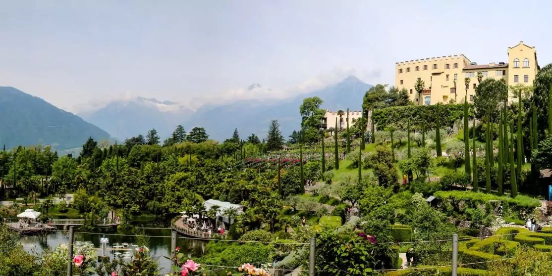 Terrassierter Garten mit Teich, Blumen und Kakteen, Menschen auf Wegen, gro&szlig;es helles Geb&auml;ude auf einem H&uuml;gel, Berge im Hintergrund