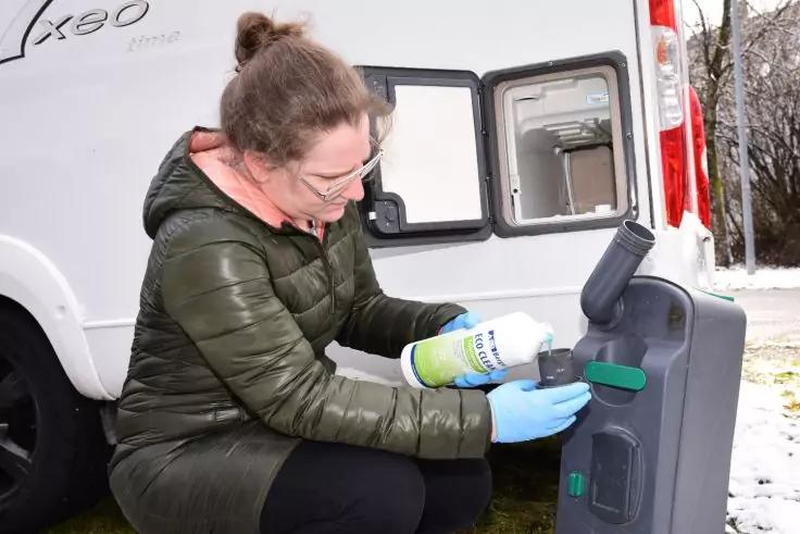 Frau kniet neben einem ausgebauten tragbaren Toilettenbeh&auml;lter, tr&auml;gt eine gr&uuml;ne Steppjacke, blaue Einmalhandschuhe und eine Brille, h&auml;lt eine Spr&uuml;hflasche in der rechten Hand und einen gr&uuml;nen Schwamm in der linken; im Hintergrund die ge&ouml;ffnete R&uuml;cks