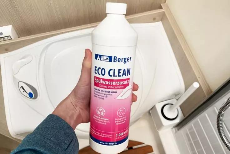 Hand mit blauem &Auml;rmel h&auml;lt eine wei&szlig;e Flasche mit pinkem Etikett Berger ECO CLEAN &uuml;ber einem kleinen wei&szlig;en Waschbecken; daneben eine wei&szlig;e B&uuml;rste in einem Halter