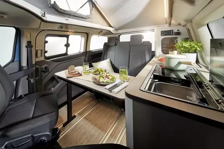 Innenraum eines Wohnmobils mit Esstisch, zwei Sitzen, Tisch mit Salat und Tellern, K&uuml;chenzeile mit Sp&uuml;le und Topfpflanze, Fenster und Dachfenster